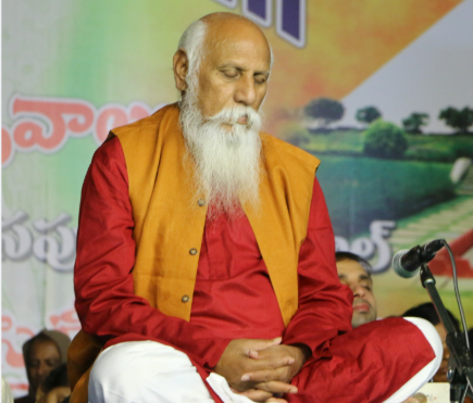 An elderly man with a white beard sitting cross-legged on a stage, eyes closed, fingers interlaced in his lap