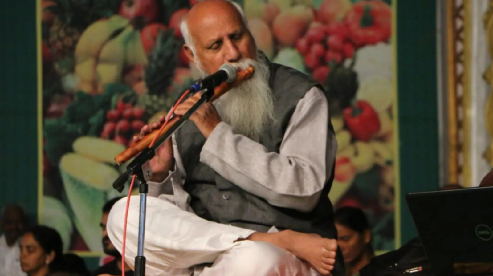 A bearded man in a grey waistcoat playing a wooden flute into a microphone